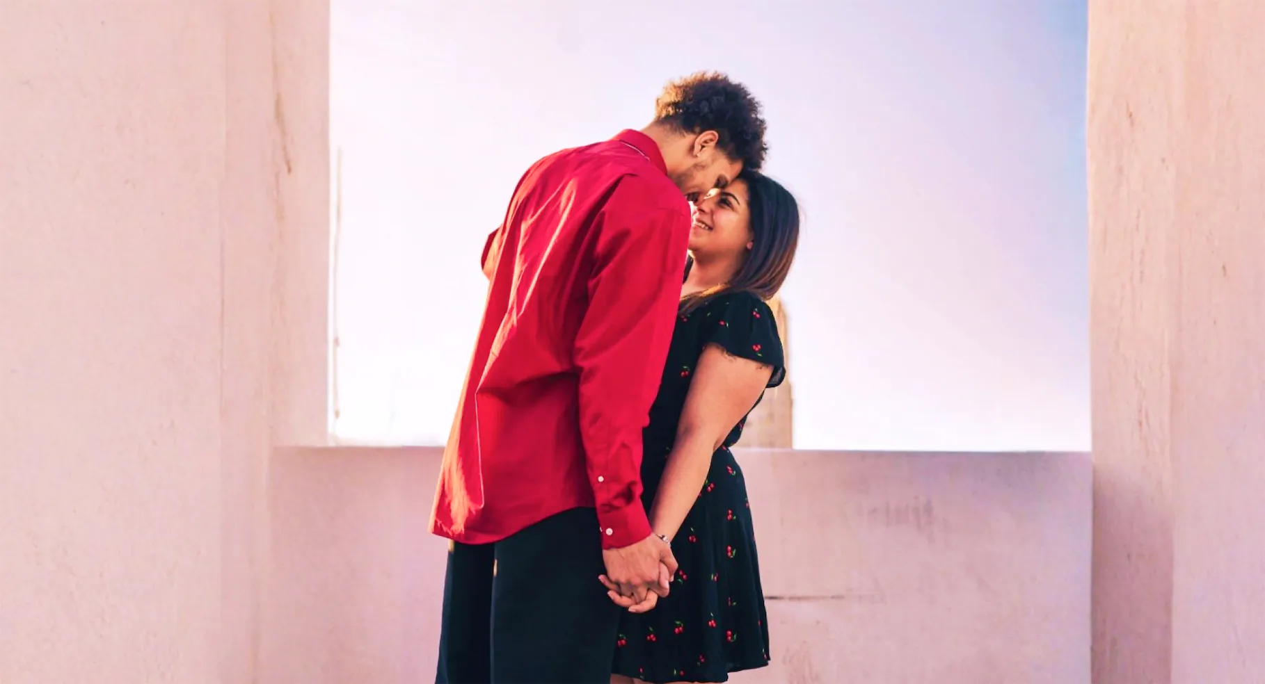 A couple stands close together, holding hands, with the person on the left wearing a red shirt and the person on the right wearing a black dress with red cherry patterns. They are looking at each other and appear to be about to kiss.